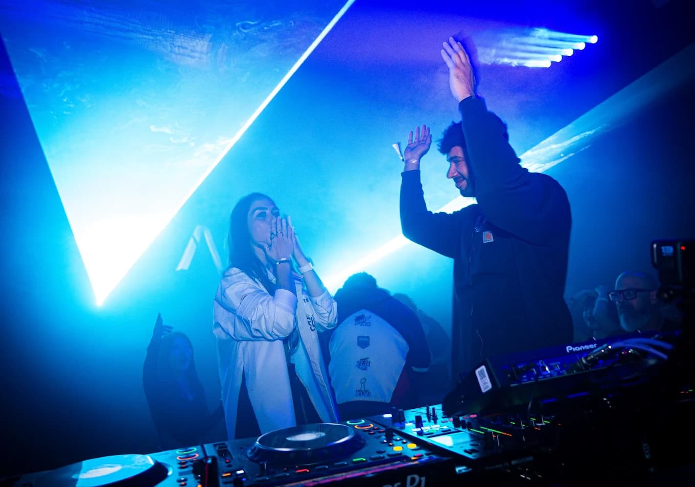 Two DJs celebrating behind the decks at 00:00 as lasers cut through the haze