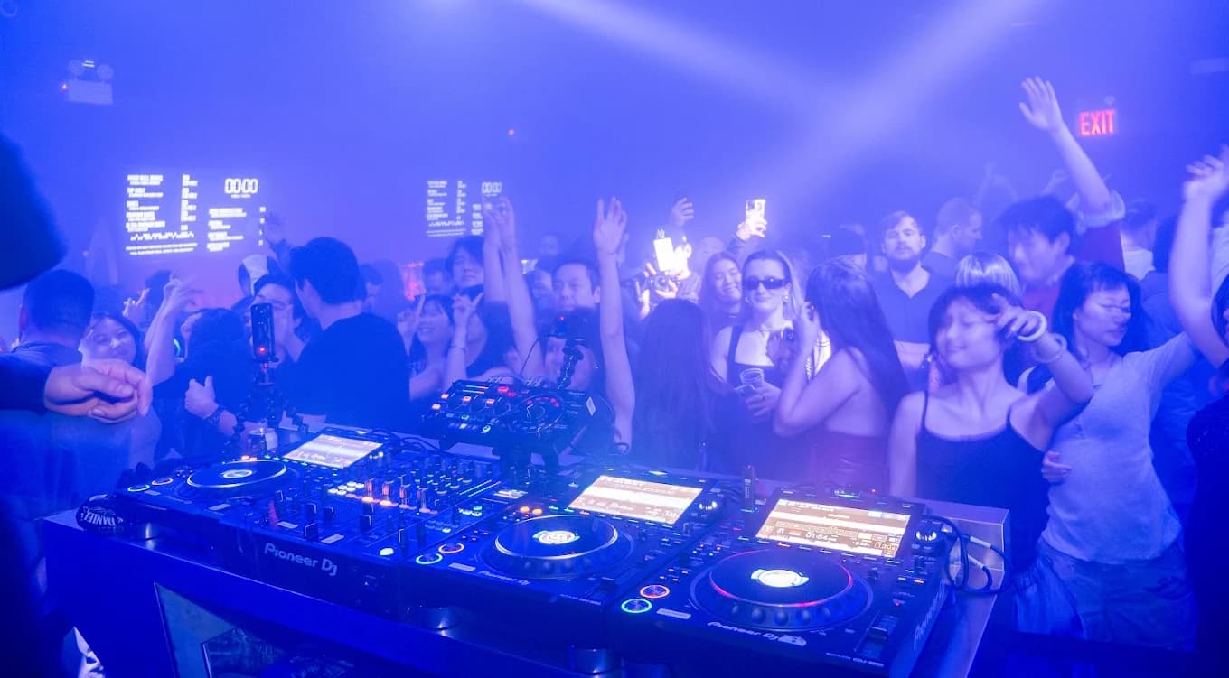 The DJ booth at 00:00 facing a crowd with hands in the air under blue lights